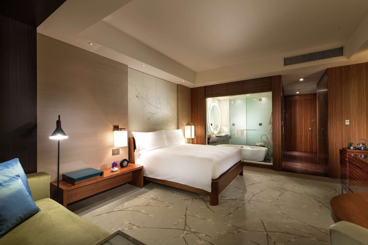 Una habitación de hotel moderna con una cama grande, mesita de noche, lámpara y vistas al baño.
