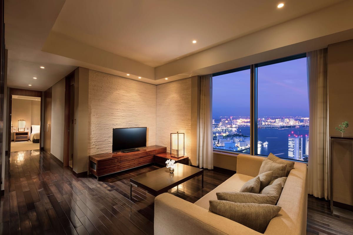 Habitación de hotel moderna con un ventanal grande que ofrece vistas a la ciudad, un televisor de pantalla plana, un mueble de entretenimiento de madera y un rincón de descanso cómodo.