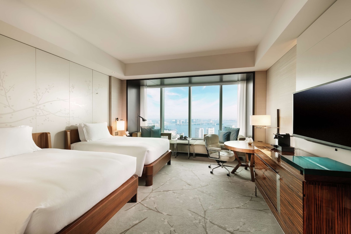 A hotel room with two beds, a desk, a chair, a television, and a large window offering a city view.