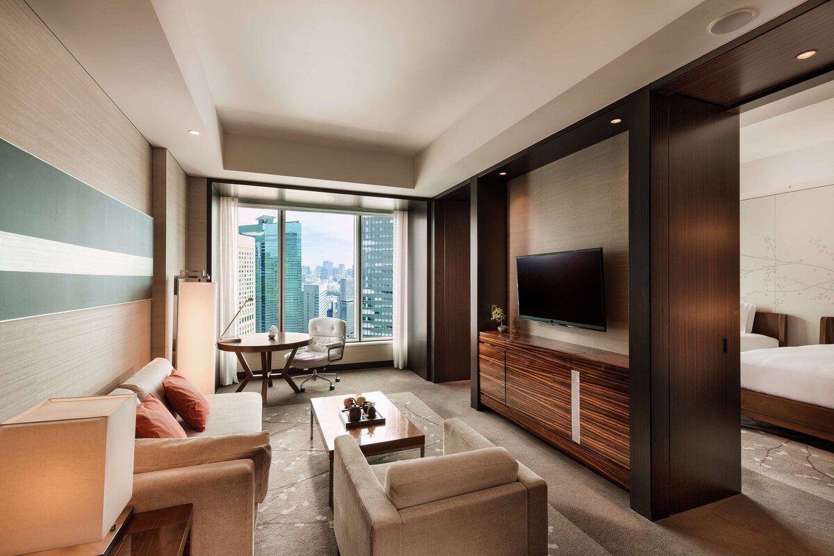 A hotel room with a large bed, a TV, a seating area with a table and chairs, and a view of the cityscape through the window.