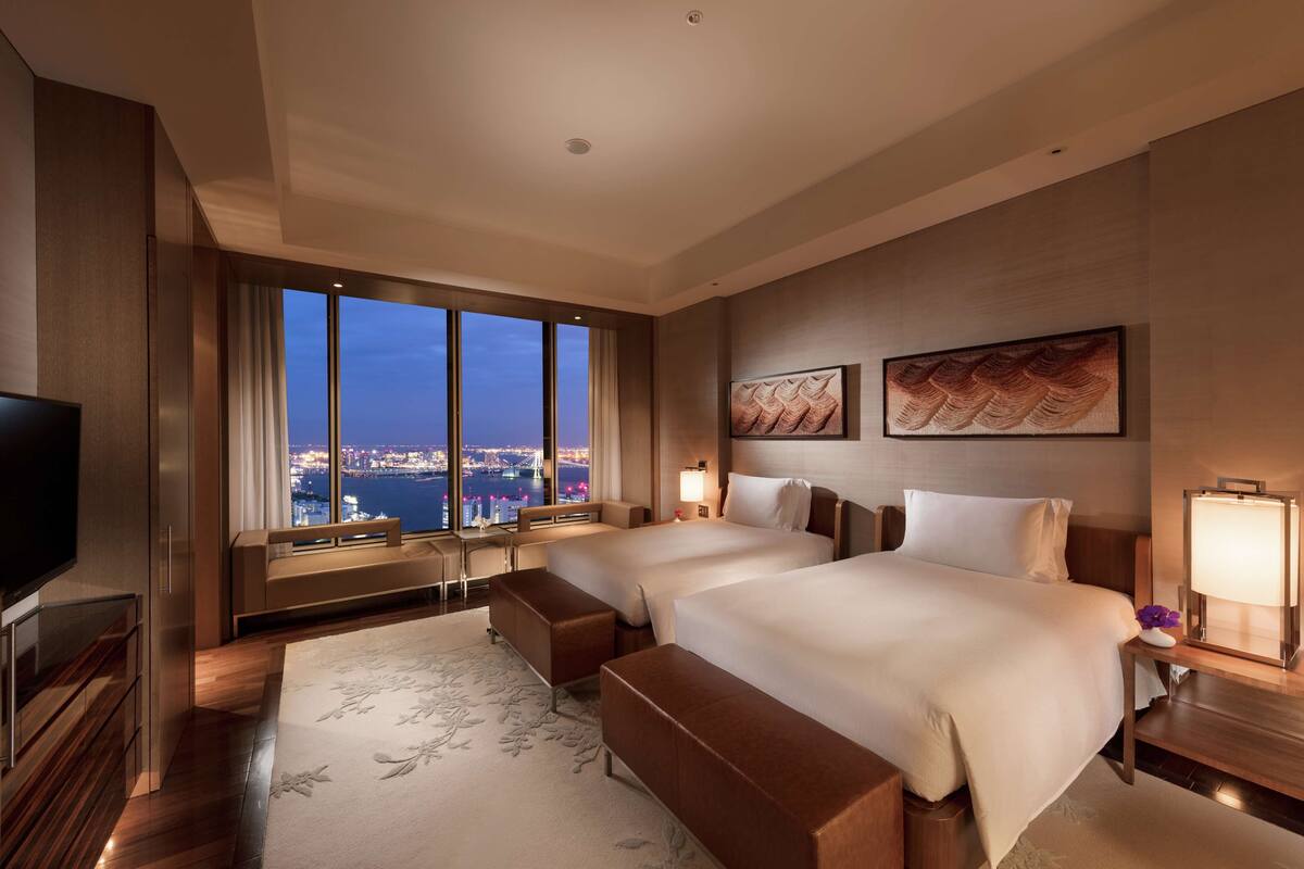 Habitación de hotel con dos camas, televisión, escritorio y una ventana grande con vista a la ciudad.