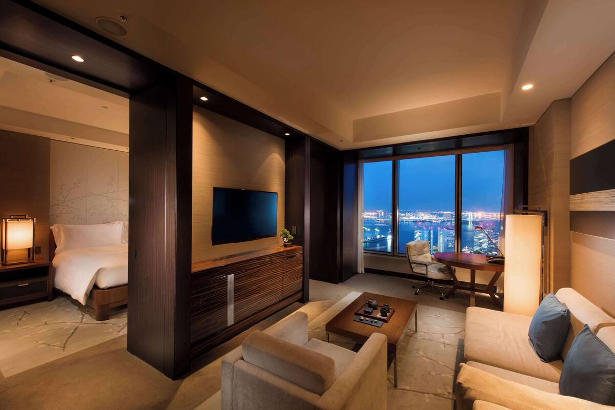 A modern hotel room with a large bed, a flat-screen TV, a seating area with a coffee table, and a city view through large windows.