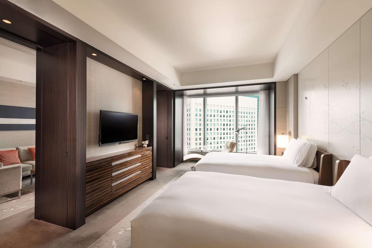 Habitación de hotel con dos camas, televisión, escritorio y ventana grande con vistas a la ciudad.