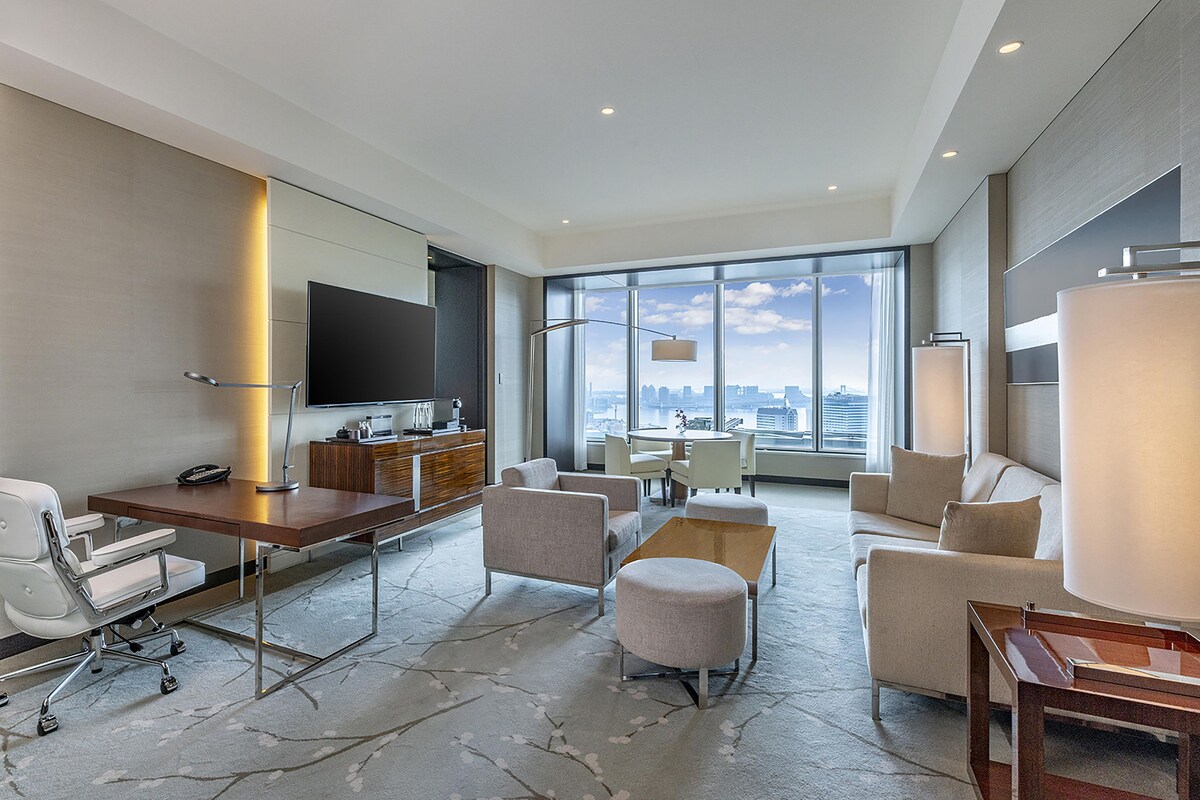 A modern hotel room with a large desk, seating area, and a city view.