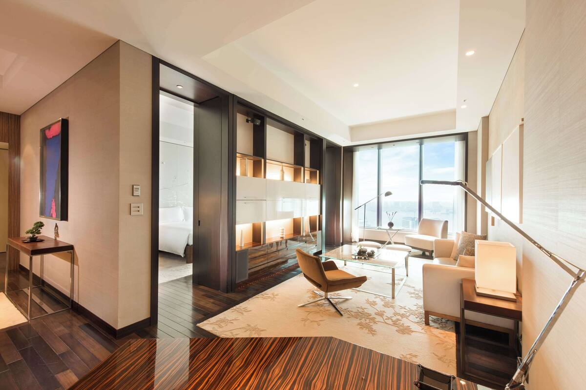 Una habitación de hotel moderna con un ventanal grande, un sofá, una mesa de centro y un escritorio con lámpara.