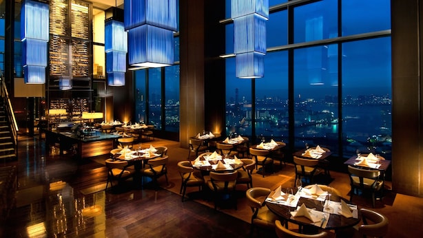 A modern restaurant with high ceilings, large windows, and blue hanging lights. Tables are set with white napkins and glasses. - Conrad Tokyo (Tokyo)