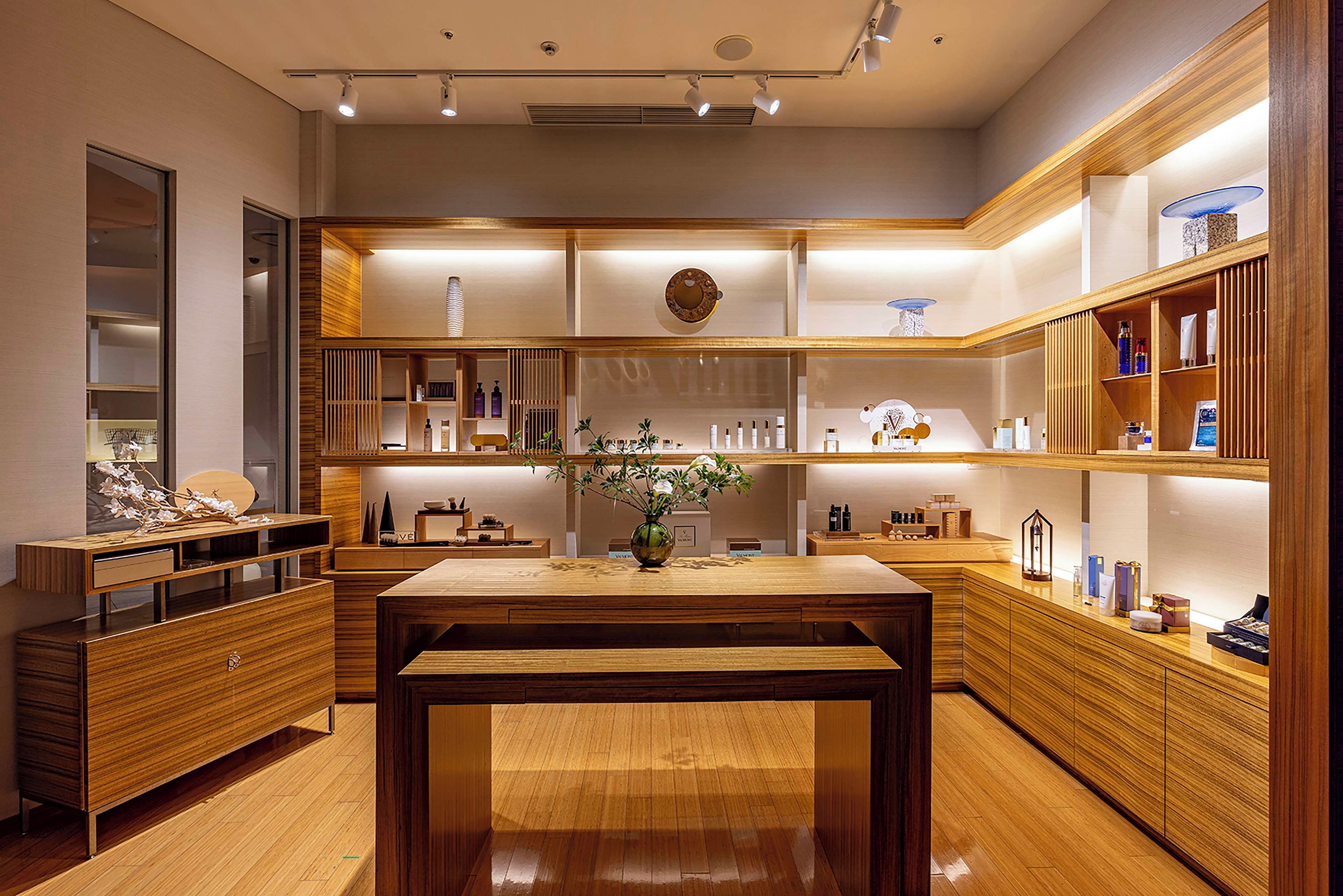 A modern hotel lobby with a wooden reception desk, shelves with toiletries, and a vase with flowers.
