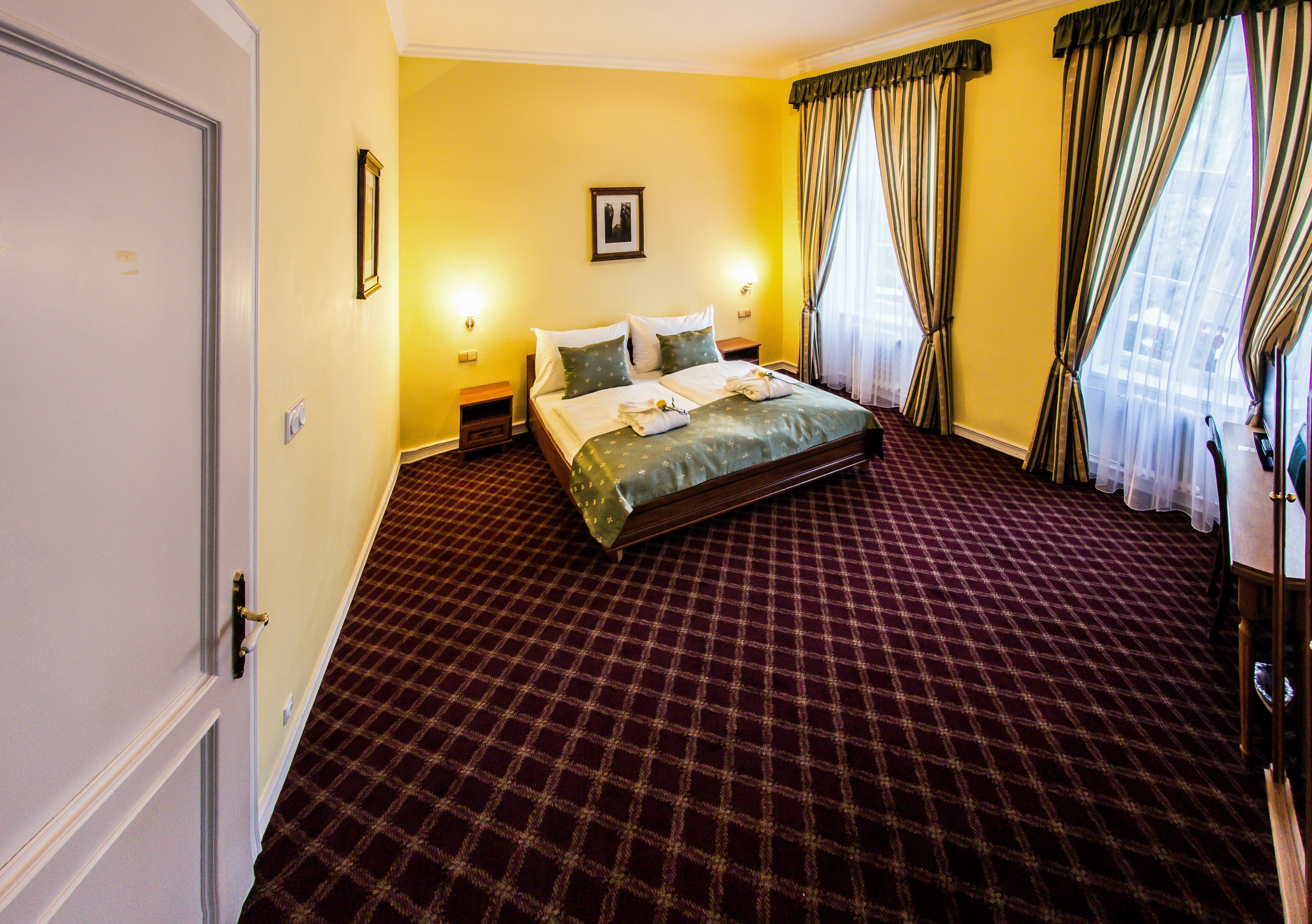 A hotel room with a checkered carpet, a bed with pillows, a nightstand, and a framed picture on the wall.