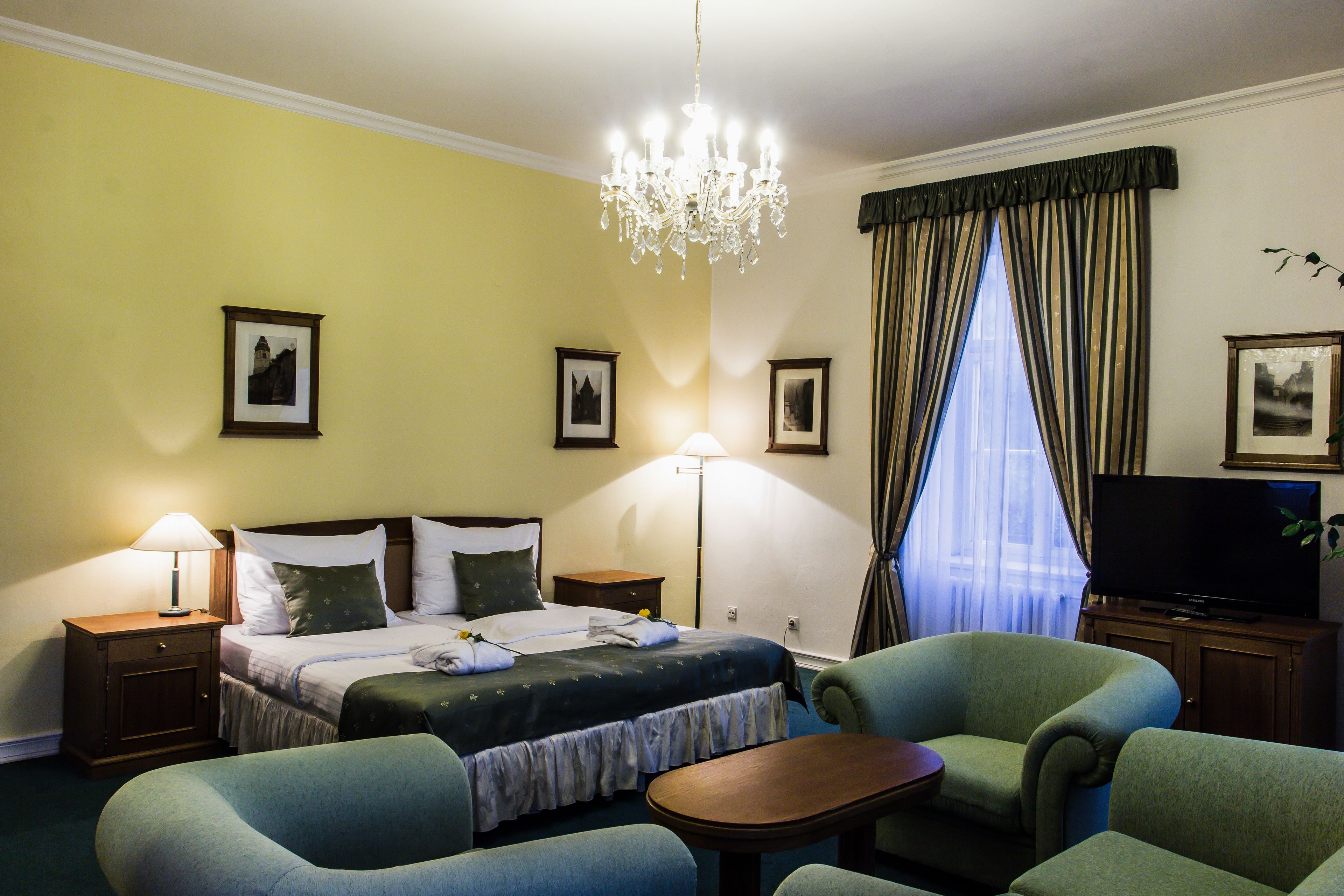 A hotel room with a bed, two armchairs, a small table, a TV, and a chandelier.