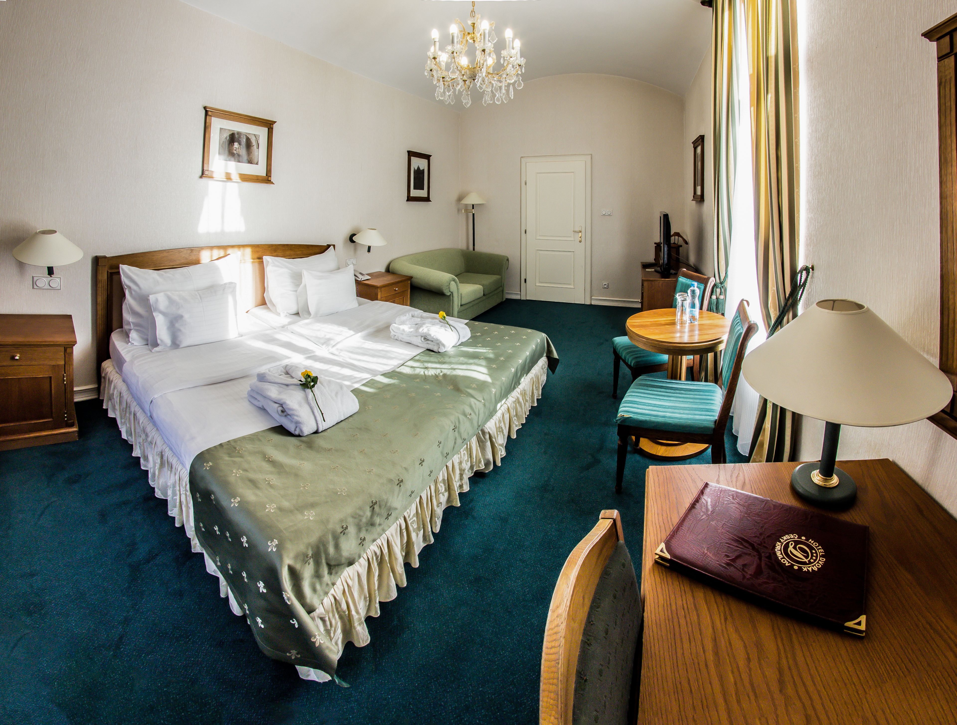 Een hotelkamer met een opgemaakt bed, een groene bank, een eettafel met stoelen, een lamp en een bijzettafeltje met een boek en een glas.