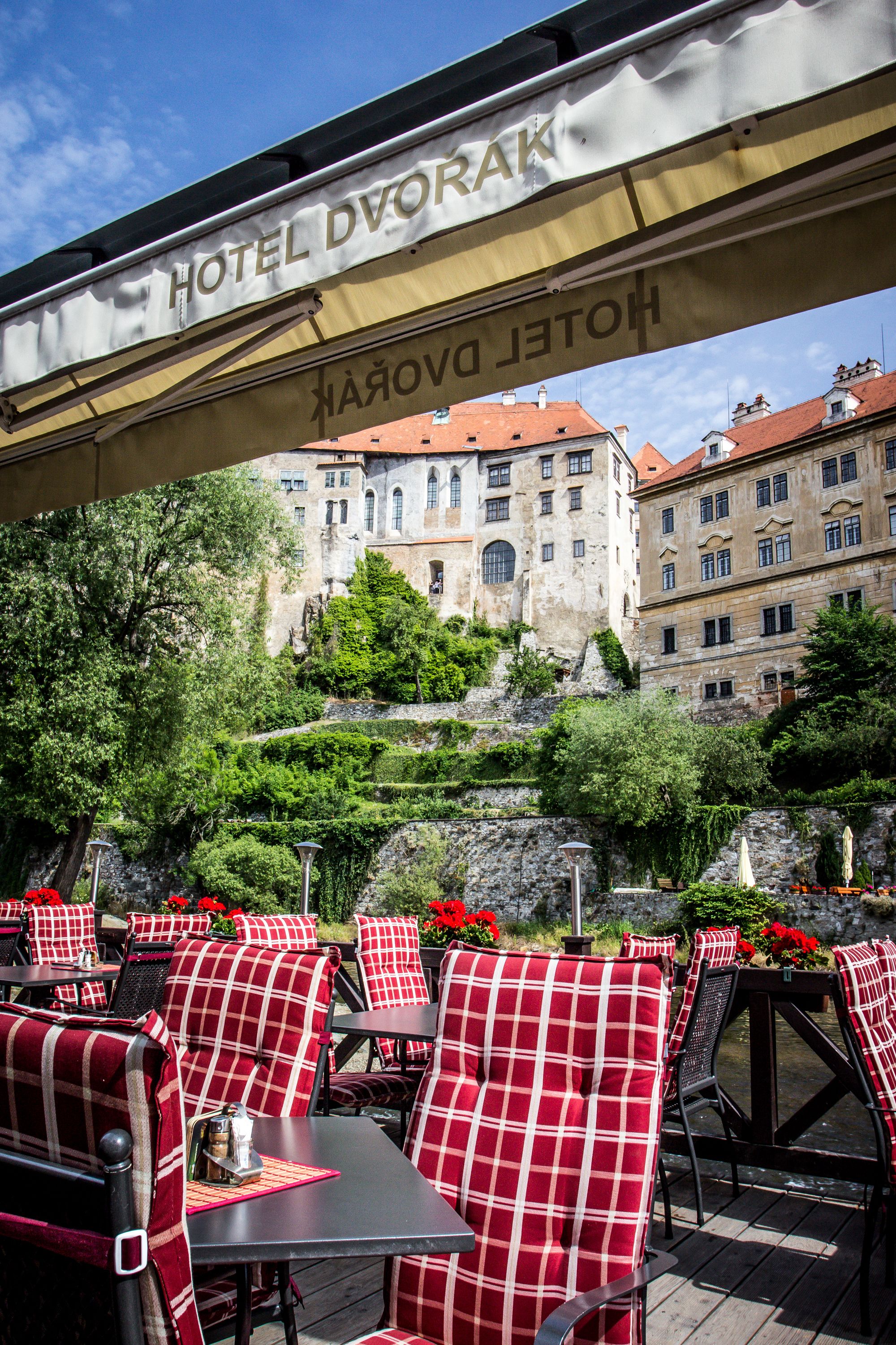 A hotel outdoor seating area with red and white checkered chairs and tables, a stone wall, and a building with red roofs in the background.