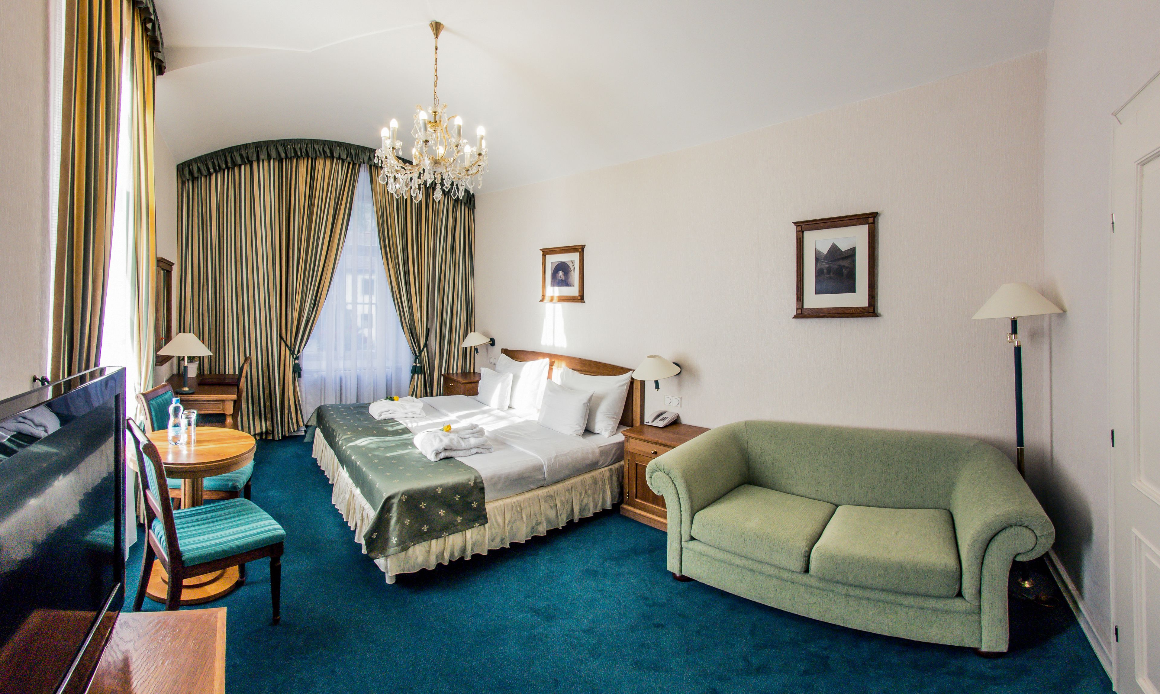 Une chambre d’hôtel avec un lit, un fauteuil vert, un bureau avec une chaise, une télévision, un lustre et deux lampes.
