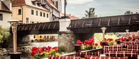 Restaurant a la riba d'un riu amb terrassa, un pont i un edifici històric amb una cúpula verda al fons.