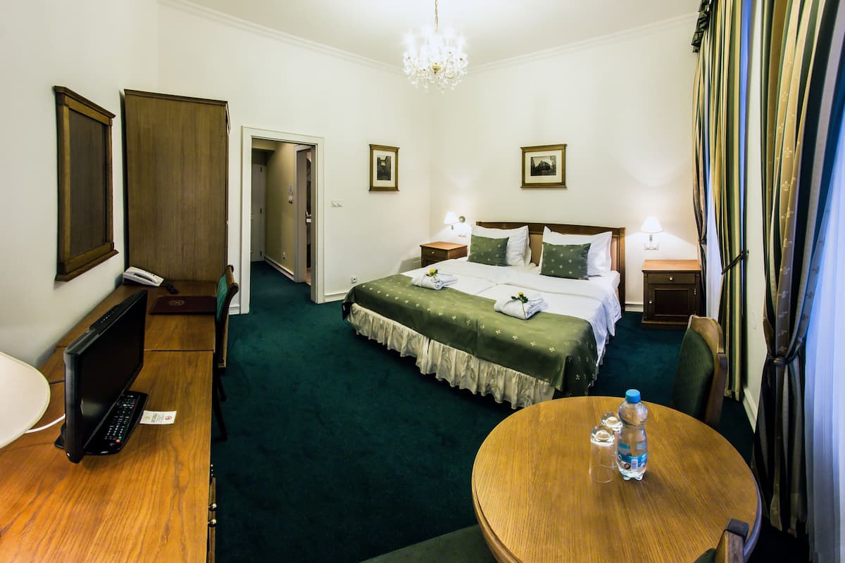 A hotel room with a bed, bedside tables, a desk with a laptop, a round table with a bottle, and a chandelier.