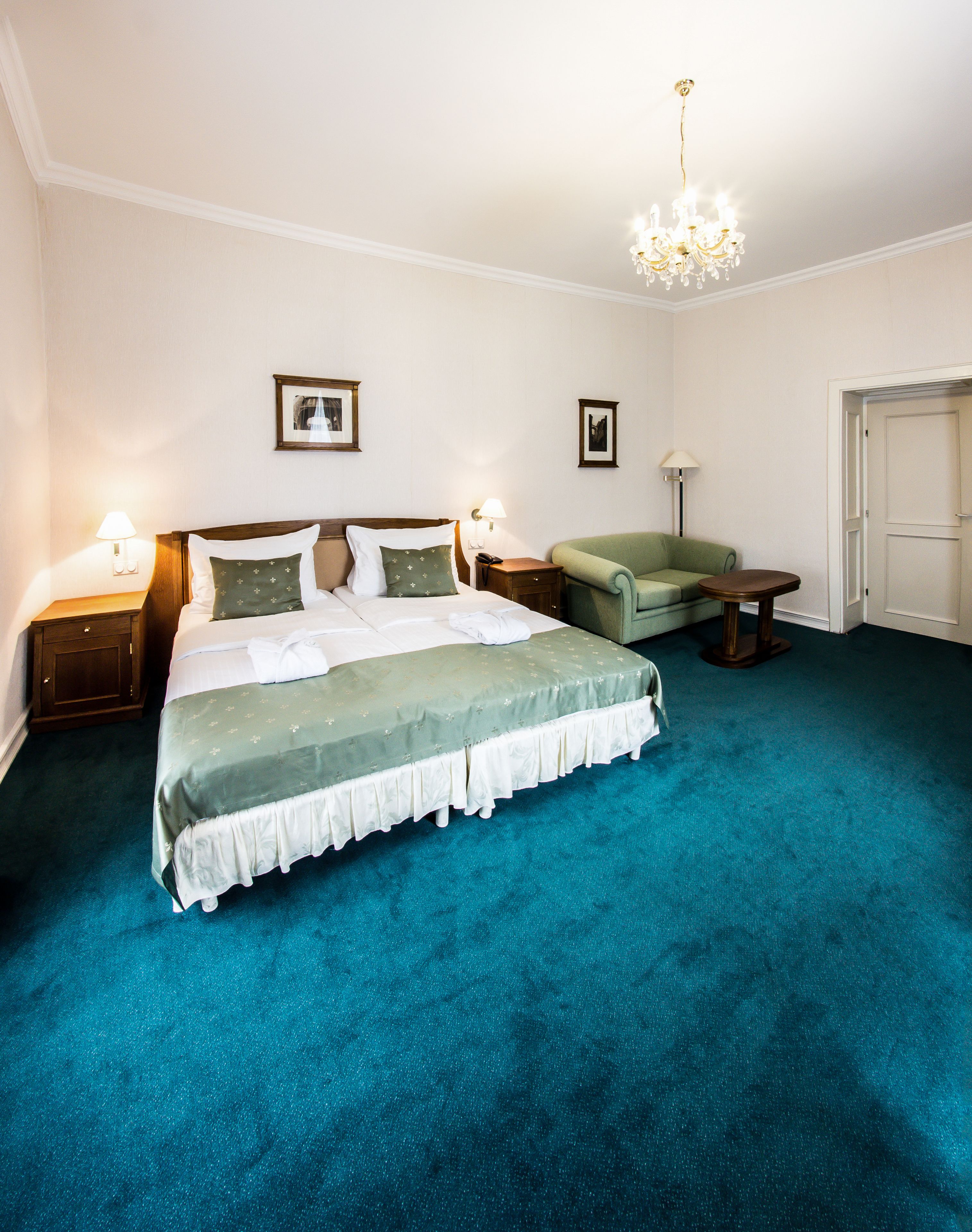 A neatly arranged bedroom with a large bed, a green armchair, a small table, and a chandelier.