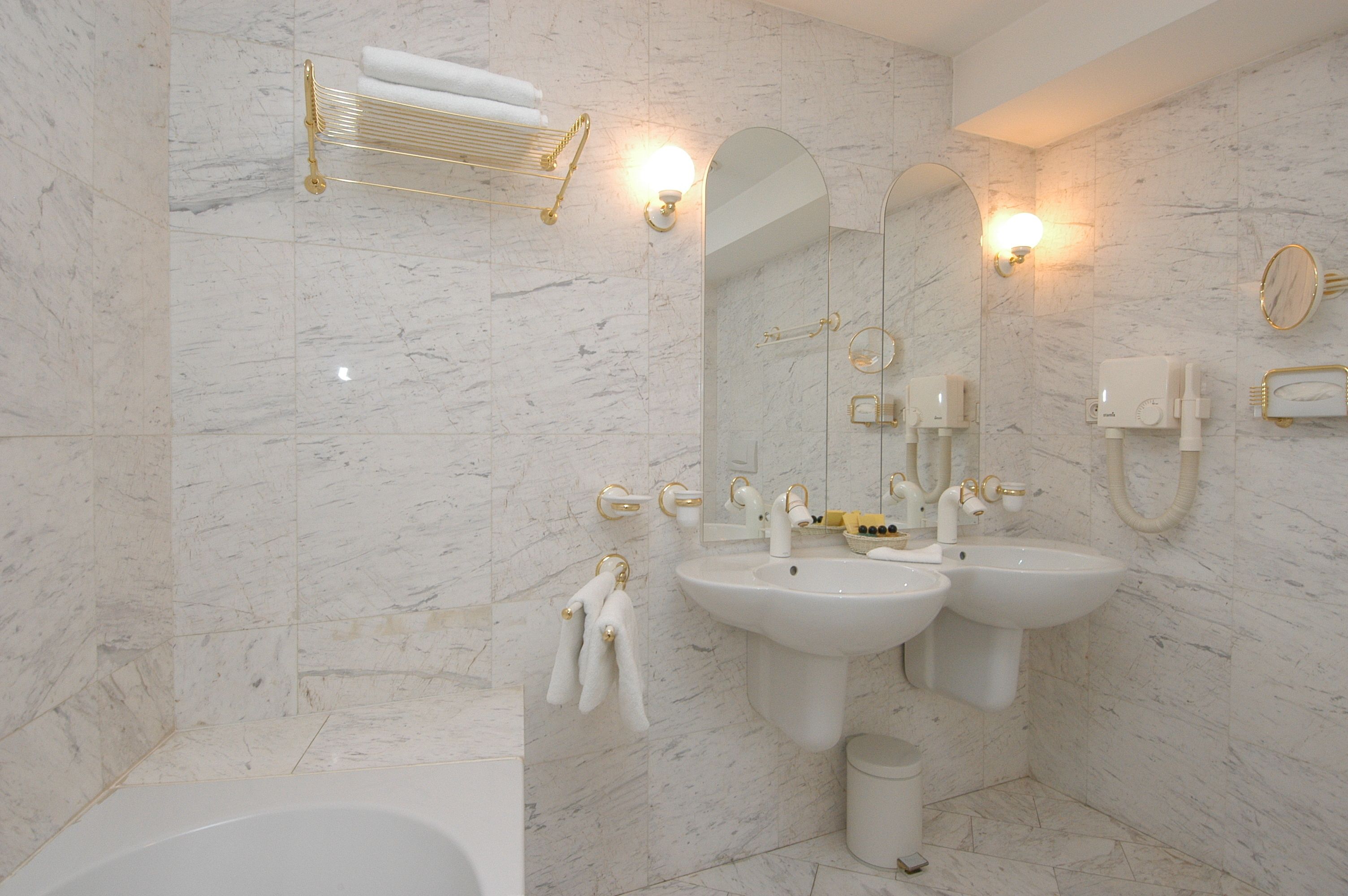 Un baño moderno con paredes de mármol, dos lavamanos, una bañera e instalaciones doradas.