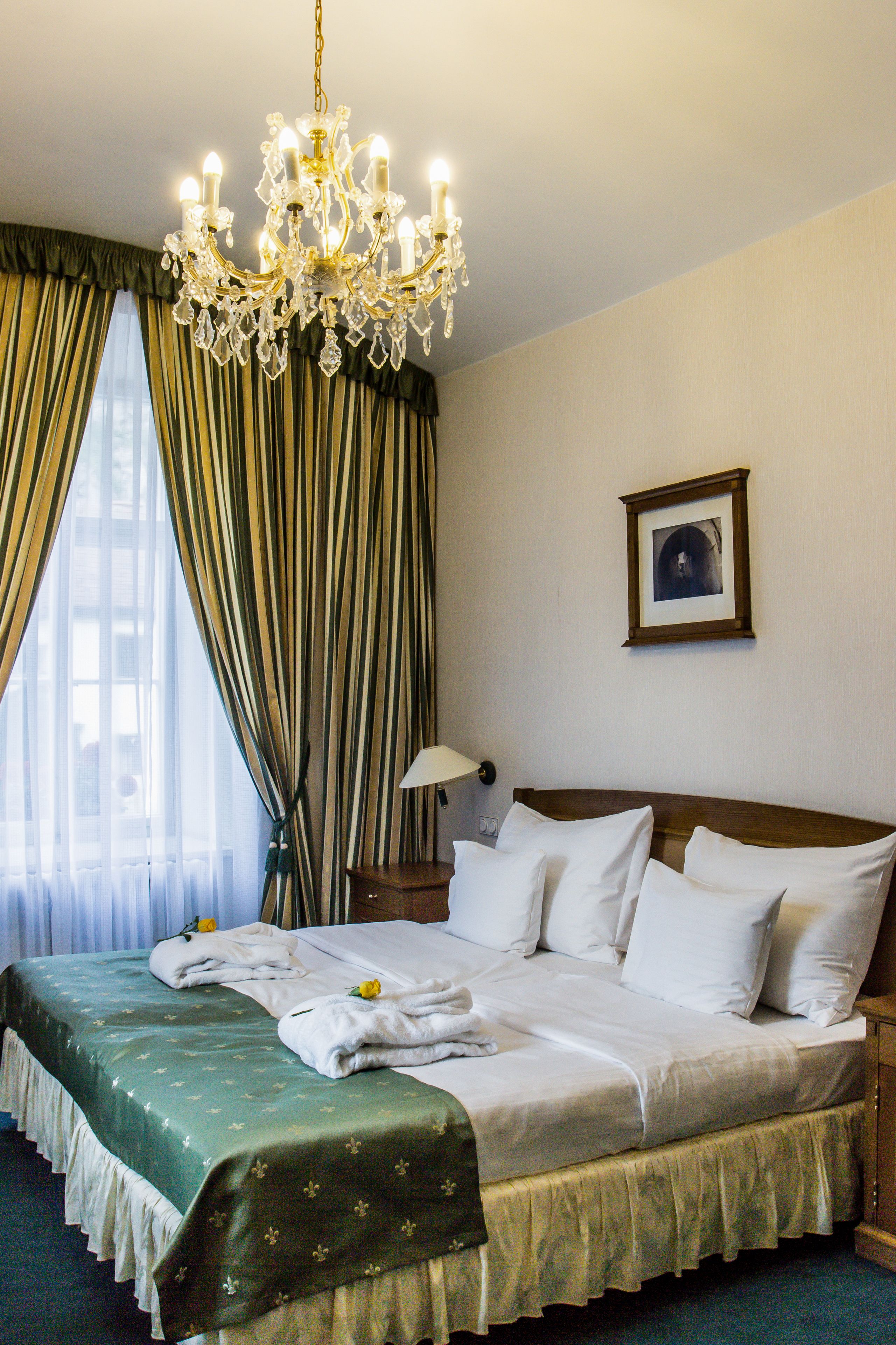 A hotel room with a large bed, a chandelier, a framed picture on the wall, and curtains on the window.