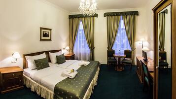 A hotel room with a large bed, a desk, two chairs, a small table, a mirror, and a chandelier.