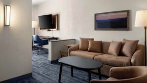 Habitación de hotel con sofá, una mesita, escritorio con computadora y silla, televisor montado en la pared y un cuadro.