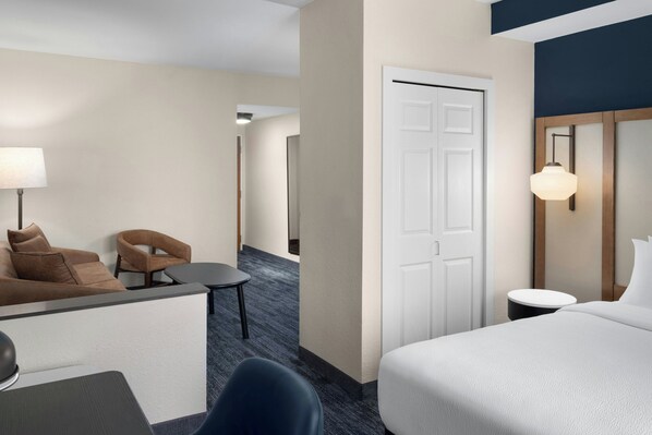 A hotel room with a bed, a desk with a lamp, two chairs, a small table, and a door.