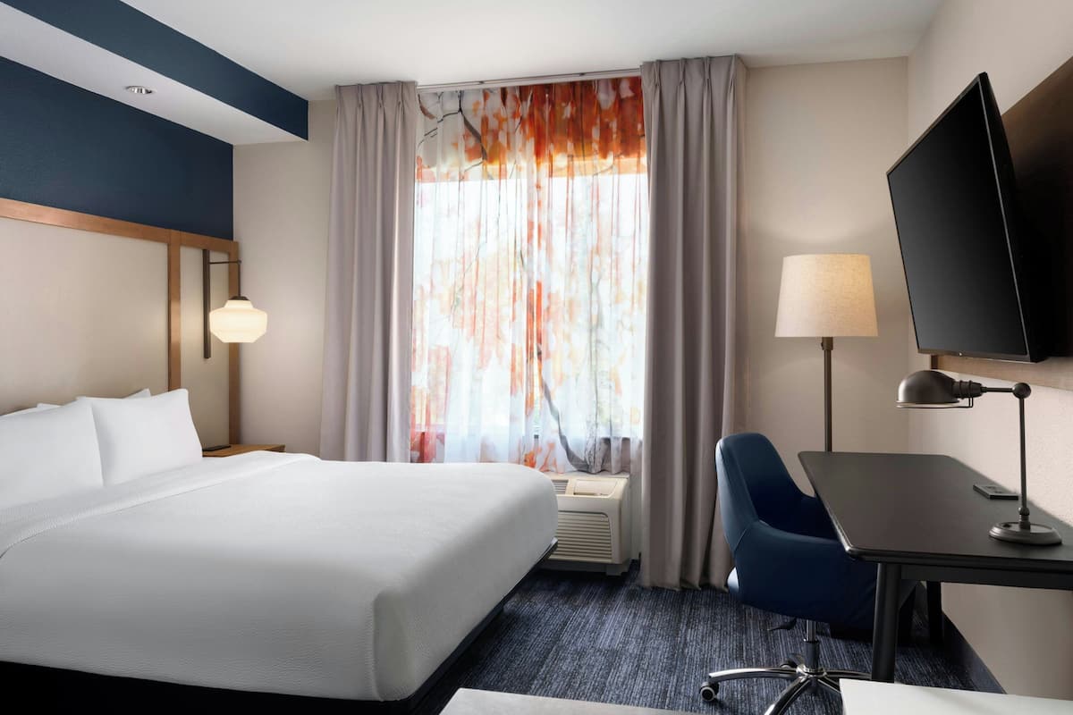 Habitación de hotel con una cama grande, un escritorio con silla, televisor de pantalla plana, lámpara y ventana con cortinas.