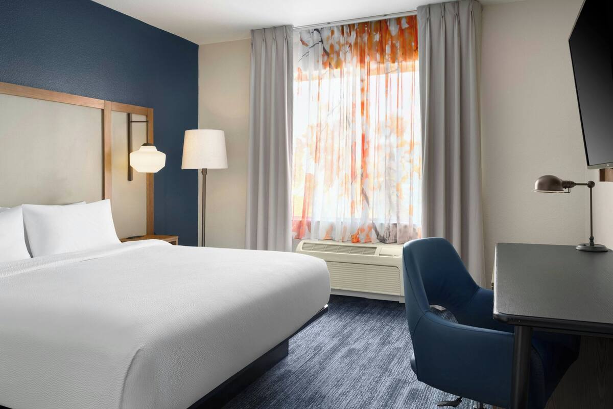 Habitación de hotel con una cama grande, un escritorio, una silla, un televisor y una ventana con cortinas.