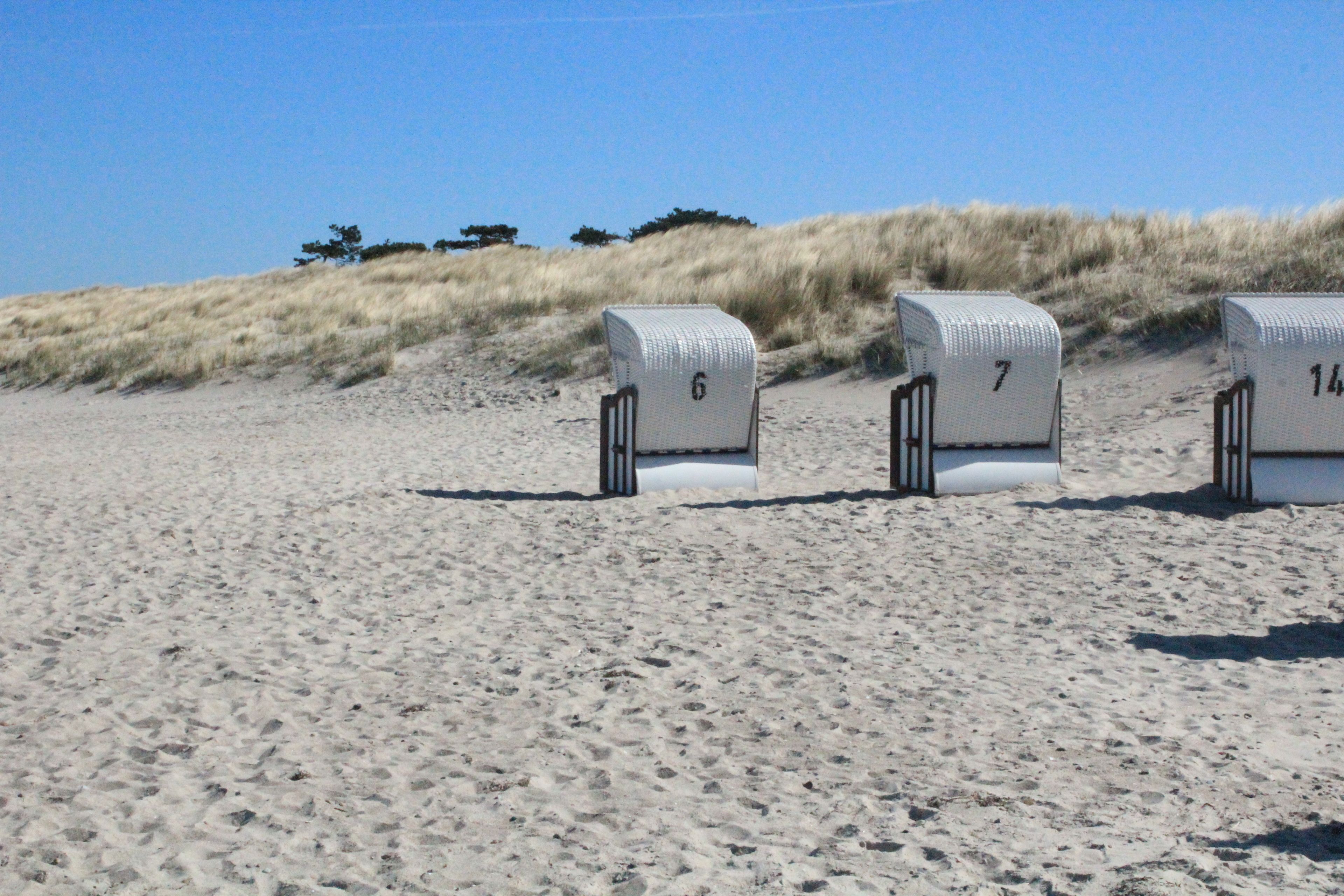 Trois chaises de plage, numérotées, sont alignées sur une plage de sable, avec des dunes en arrière-plan.