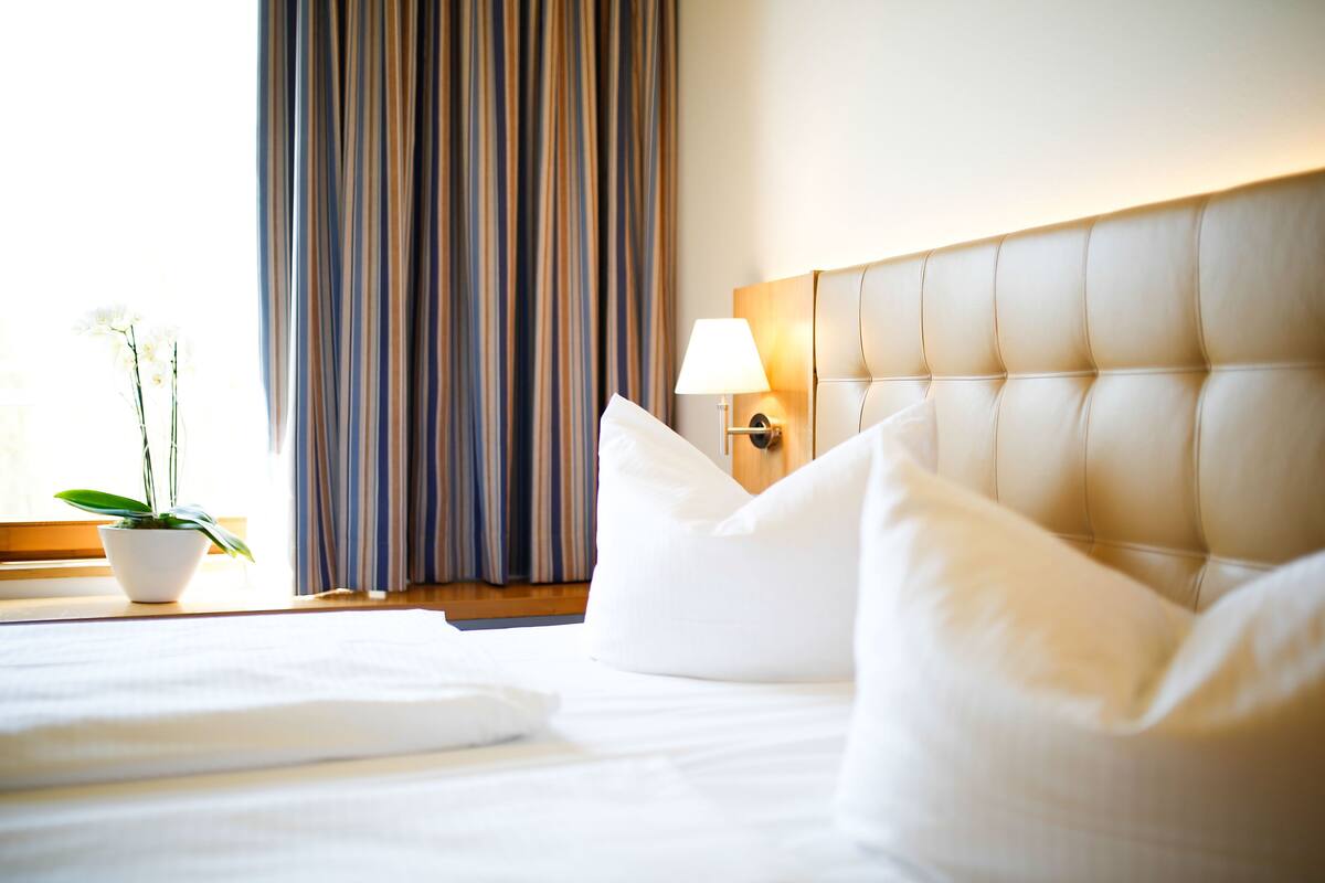 A hotel room with a neatly made bed, a bedside lamp, a potted plant, and a striped curtain.