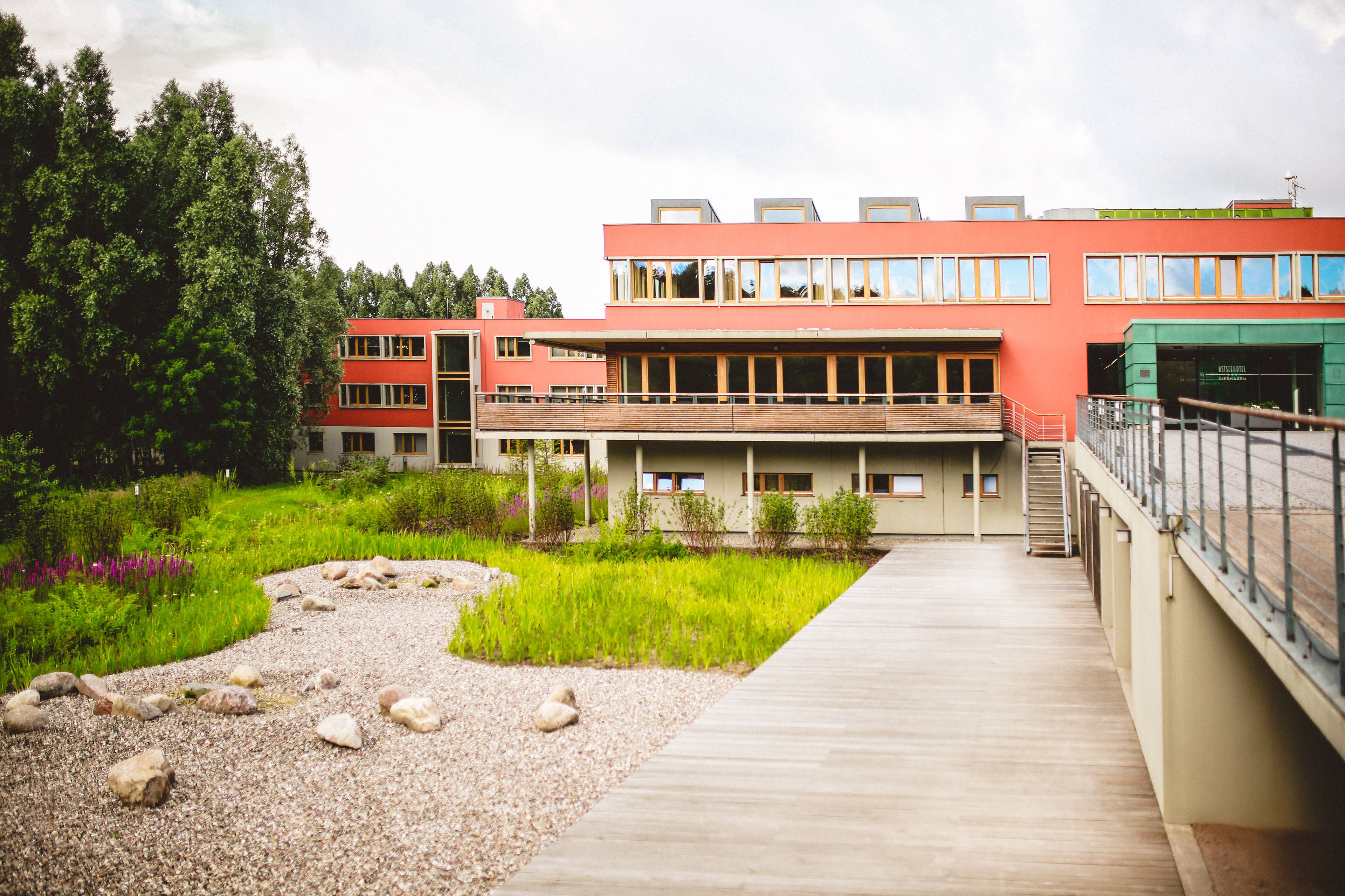 Ein modernes Gebäude mit roter Fassade, einer Holzterrasse und einem Gartenbereich mit Steinen und Pflanzen.