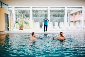 An indoor swimming pool with clear glass walls, a person in a blue shirt standing in the background, and two people swimming in the pool. - Ostseehotel Dierhagen (Ostseebad Dierhagen)