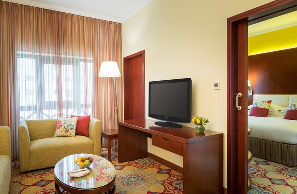 A hotel room with a sofa, a desk with a television, a small table with a fruit bowl, and a bed with a patterned bedspread.