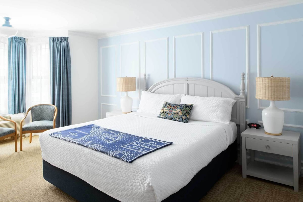 Habitación de hotel ordenada con una cama grande, dos mesitas de noche, una silla y una ventana con cortinas.