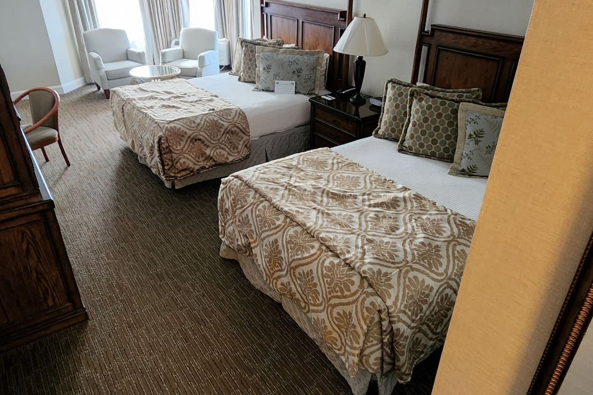 A hotel room with two beds, a desk, a chair, and a lamp.