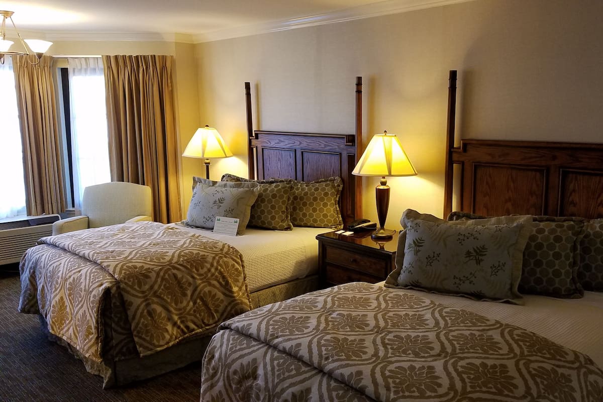 A hotel room with two beds, each with patterned bedding, two bedside tables with lamps, a chair, and a window with curtains.