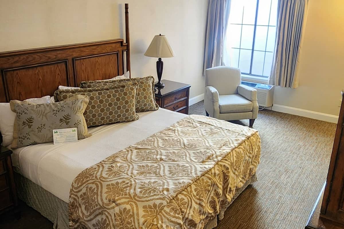 A hotel room with a large bed, a bedside table with a lamp, a chair, and a window with curtains.