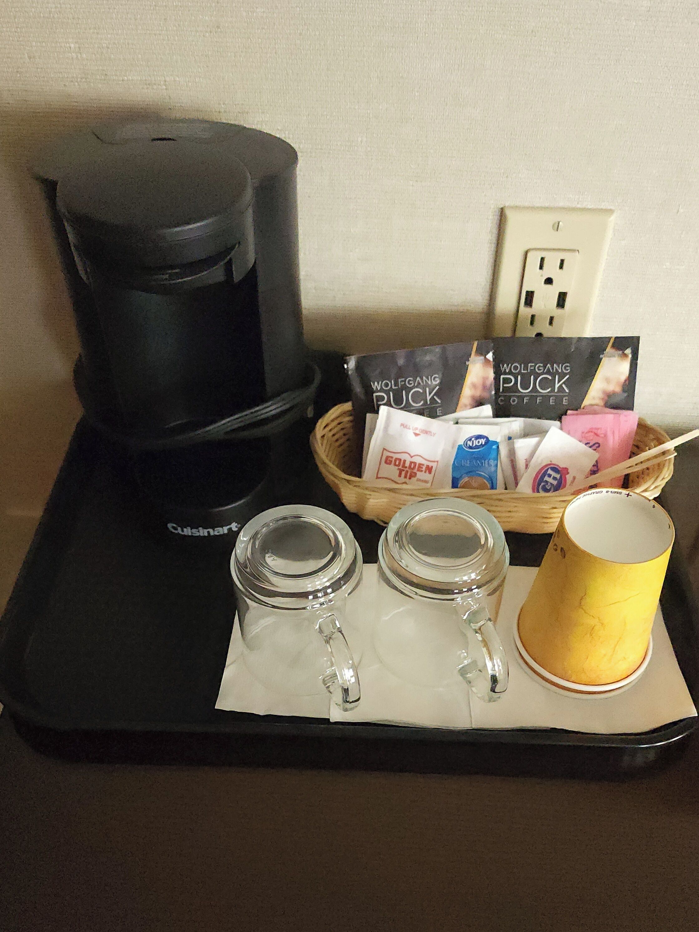 A coffee maker, a basket with coffee packets, and two mugs on a tray.