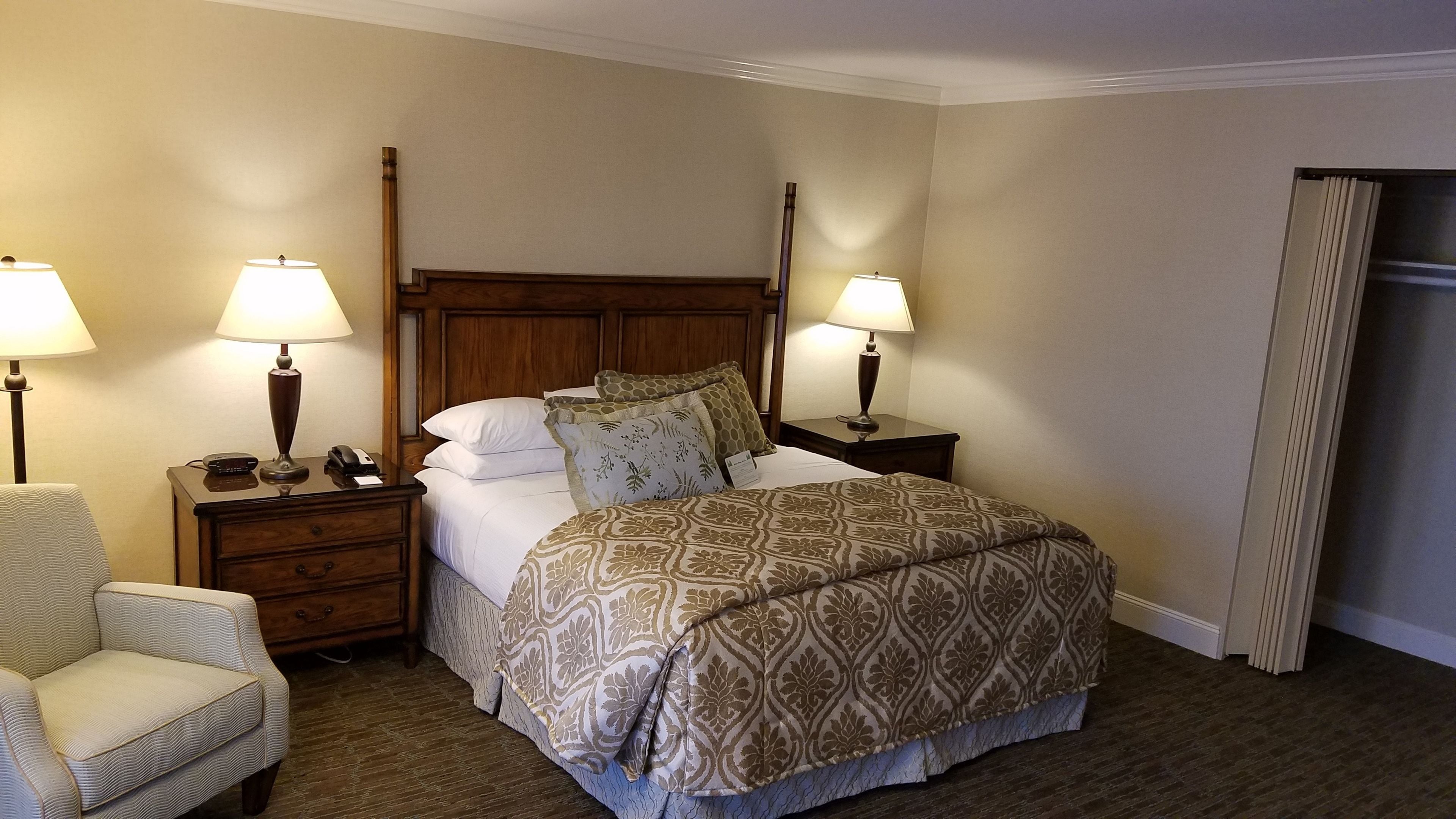 Ein Hotelzimmer mit einem hölzernen Kopfteil, zwei Nachttischen mit Lampen, einem Bett mit gemusterten Bettwäsche, einem Sessel und einem Kleiderschrank.