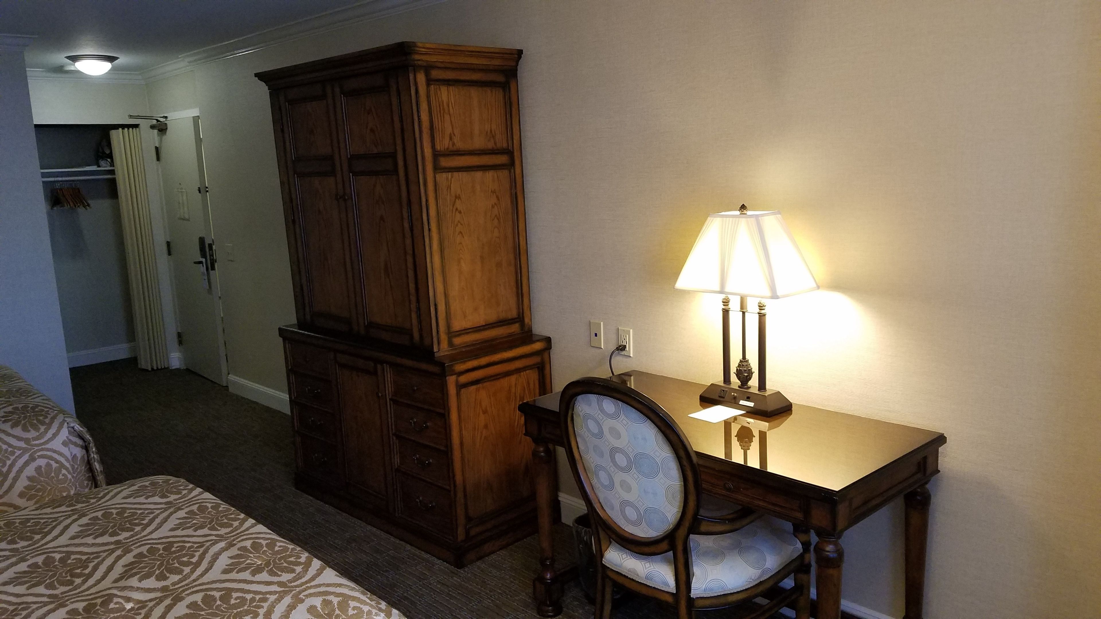 Ein Hotelzimmer mit einem Holzkabinett, einem Schreibtisch mit Lampe, einem Sessel und einem Bett mit gemusterten Betttextilien.