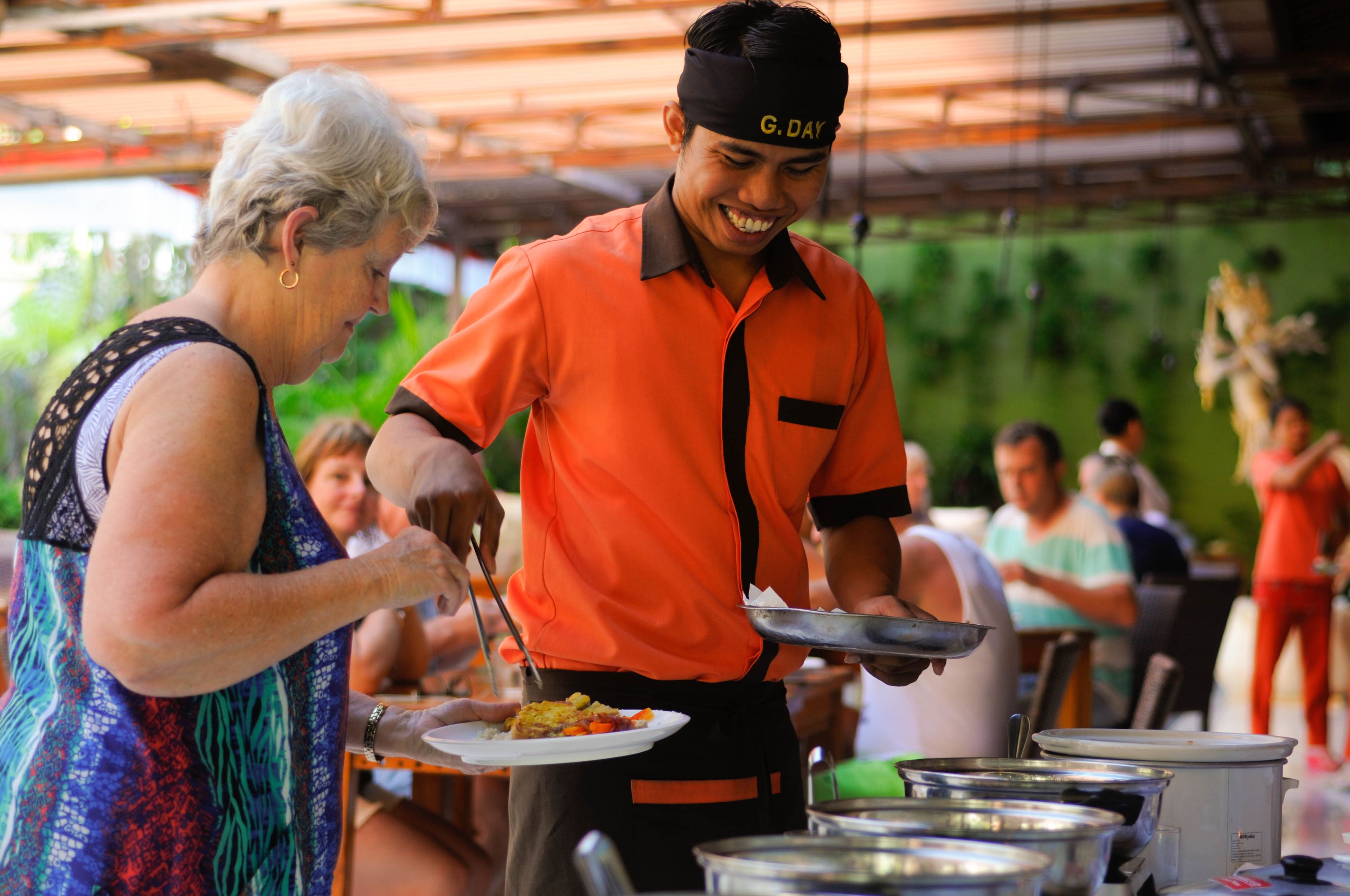 A person serving food at an outdoor event with a covered area and other attendees in the background.