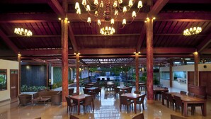 Un vaste espace de restauration intérieur, doté de tables et de chaises en bois, d’un lustre central et offrant une vue sur un jardin extérieur.