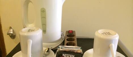 Un coin café comprenant une bouilloire électrique blanche, deux tasses blanches, une cuillère et une dosette de café Nescafé.