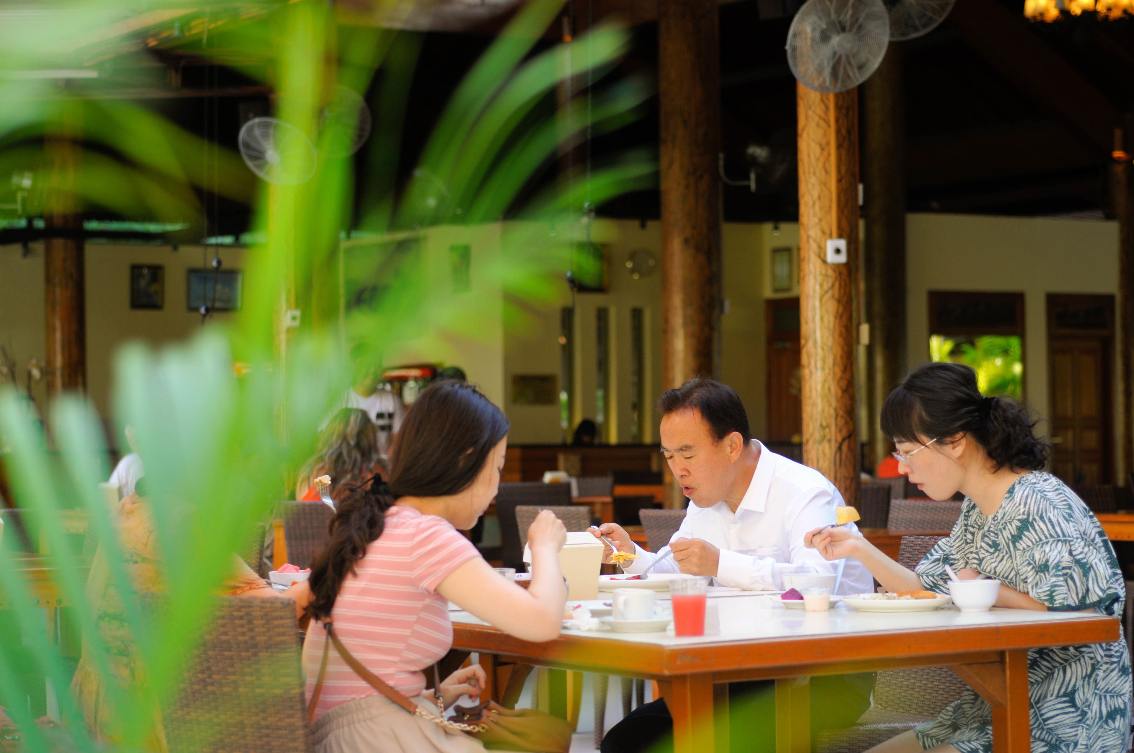 A group of people dining at an outdoor restaurant with a wooden table and chairs, surrounded by greenery.