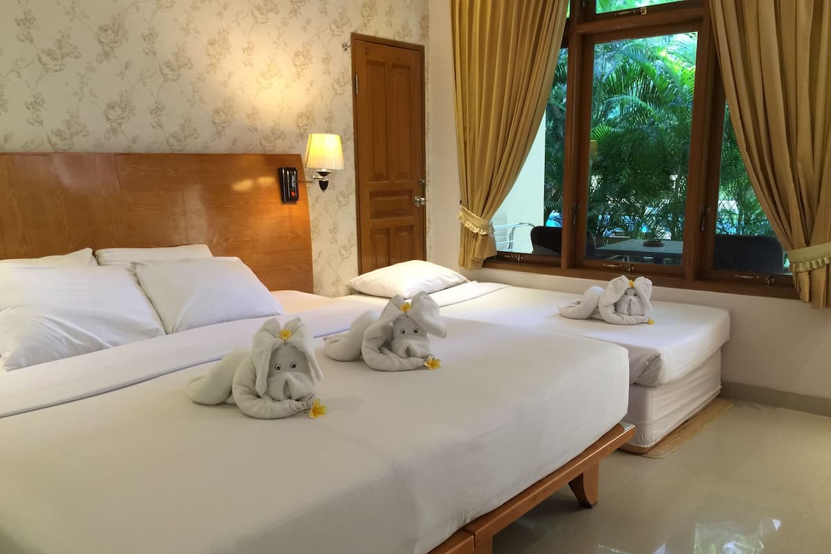 Quarto de hotel com duas camas, cada uma com lençóis brancos e cabeceira de madeira. Há duas toalhas macias em formato de elefante sobre as camas. Uma luminária está fixada na parede ao lado da porta, e uma janela com cortinas é visível.