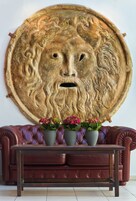 Uma obra de arte redonda e texturizada com o rosto de um leão, colocada acima de um sofá de couro marrom escuro com duas plantas em vasos.