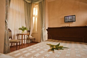 Quarto de hotel com uma cama, uma mesinha com um vaso de flores, uma cadeira e uma televisão na parede.