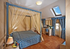 A bedroom with a bed, a desk, a chair, and a small table with a lamp and fruit bowl.