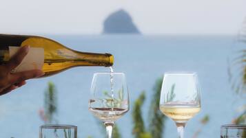 A table with two wine glasses, a bottle of wine, and a plate of food by a window overlooking the sea.