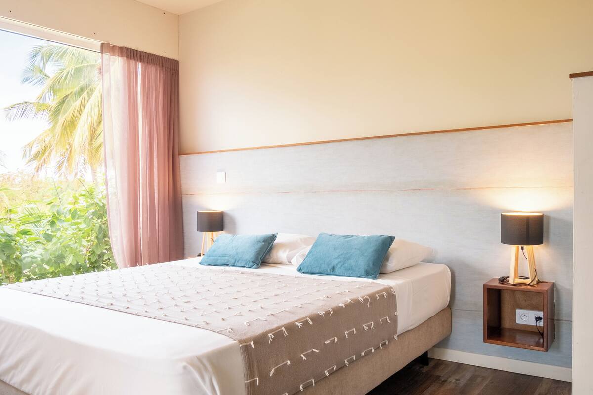 A hotel room with a large bed, two bedside lamps, a nightstand, and a view of greenery outside.