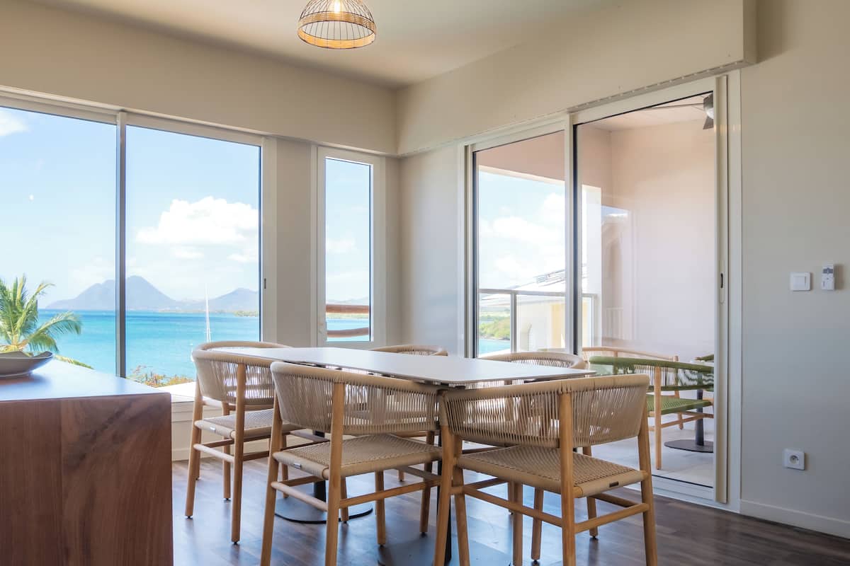 A dining area with a table and chairs, large windows with a view of the ocean and mountains, and a sliding glass door leading to a balcony.