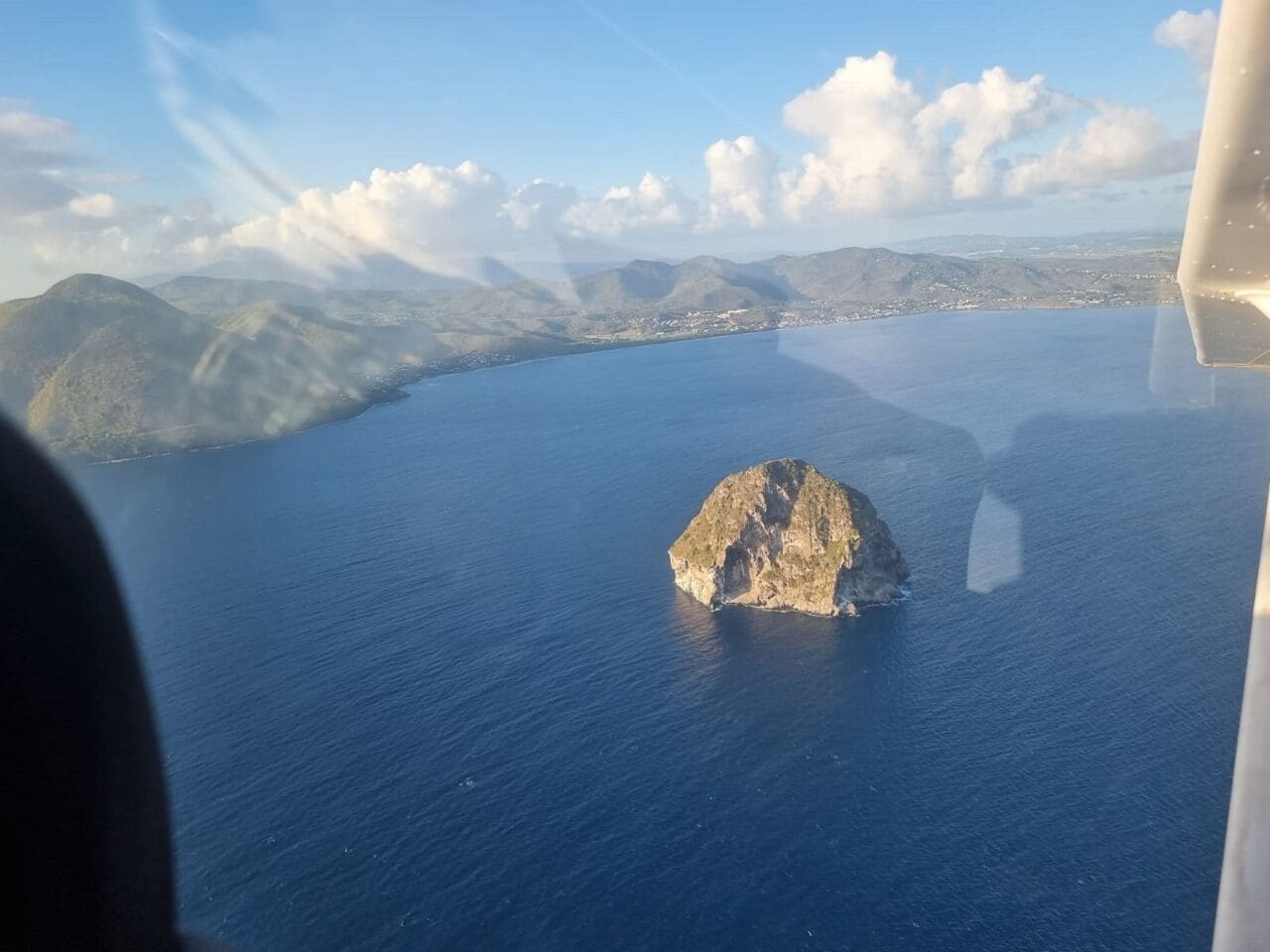 An aerial view of a coastal landscape with a prominent rocky island in the center, surrounded by calm blue waters and distant mountains.